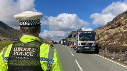 BBC Tow trucks at Pen y Pass A4086, Nant Peris, Caernarfon, Gwynedd in April 2023 with a police officer in the forefront, dressed in uniform and facing away from the camera 