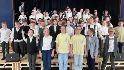 A large group of children stand on a stage in a hall. Some are casually dressed and others are wearing old fashioned clothing, waistcoats and hats. There are around 50 people in the picture all looking at the camera.