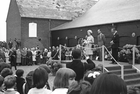 Benjamin Britten, Elizabeth II and Prince Philip stand on a platform as the late queen addresses people. Snape Maltings Concert Hall is behind them.