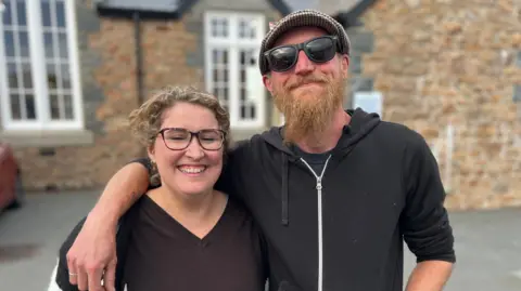 Emma Joy and her husband they are both wearing dark coloured tops. Her husband has a ginger beard and sunglasses. Emma is smiling and has blonde hair. 