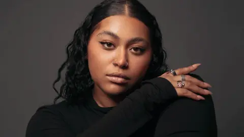 A headshot of Tegan Browne-Winter. She is wearing black, she is staring down the camera and she is wearing rings on her right hand which is resting on her left shoulder.