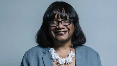 PA Media Diane Abbott smiling, is wearing a blue jacket and white flowery necklace and standing in front of a blue background