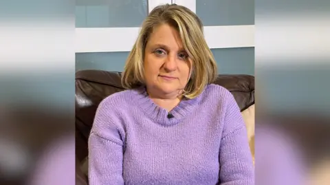 Carole Gould wearing a purple knitted jumper and sitting on a brown leather sofa in front of a blue panelled wall. She has shoulder-length blonde hair and is looking at the camera with a sad expression.