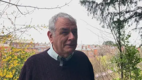 A grey haired man in a blue jumper in front of trees , and a prison building in the background 