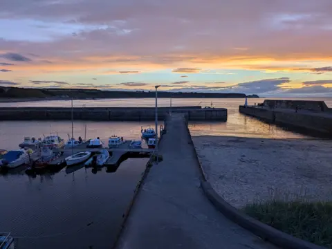 Steven McDonald A small harbour at sunset or early twilight, with tranquil water, moored boats, and a colourful evening sky of pinks and oranges.