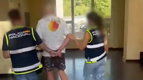 Spanish National Police Two police officers escort a person with their hands behind their back into a building