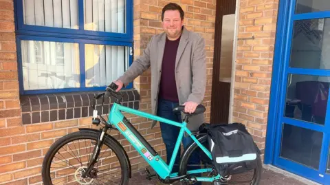 Seb Slater has short brown hair and facial hair. He is wearing a grey brown coat with a maroon t-shirt, and is standing in front of a brick building holding a bike. It is blue and has the Shrewsbury Moves logo on it.