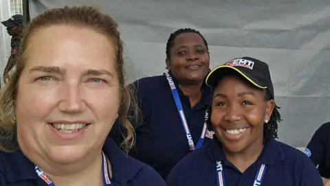 Wendy smiles for a group selife with two other women in her team. They wear the blue shirts and staff tags of the UK Emergency Medical Team.