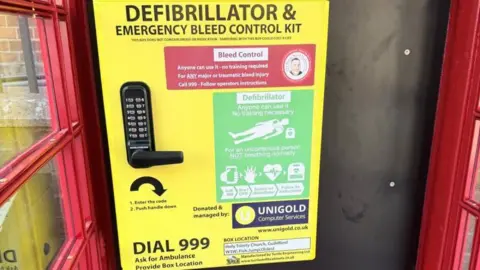 Matt Gaskin The inside of a red phone box door showing a large yellow instruction panel for a defibrillator and emergency bleed control kit, alongside a keypad lock and instructions to call 999 for access.