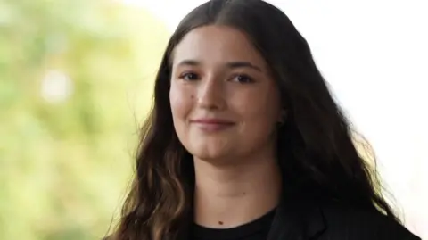 Oksana Lohvynenko A brunette girl stood in front of a nature backdrop, she smiles towards the camera as her hair flows in the wind. She is wearing a pin-striped black blazer with a rose attached to her side. 