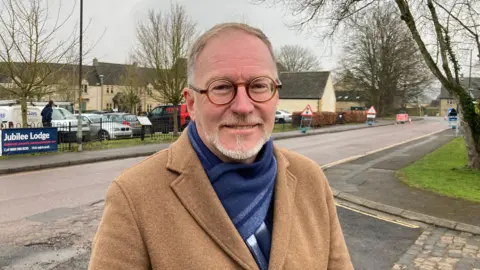 County councillor Paul Hodgkinson stands next to a road, with several buildings and a carpark behind him. 