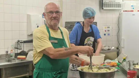 Ian White with glasses, a yellow t-shirt and green apron, preparing mashed potatoes in a large bowl while a woman with a blue hairnet, blue t-shirt and blue apron works nearby in a kitchen.