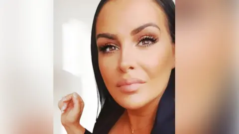 A woman with long black hair takes a selfie while standing next to a white wall.