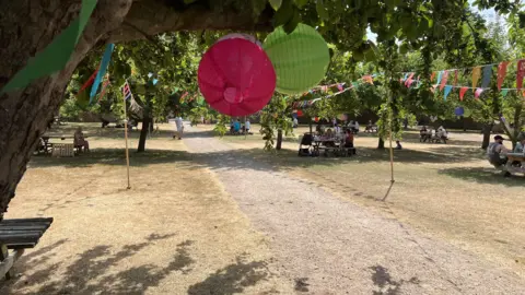 A large field that has brown short grass, with a stone path down the middle. there are trees with green leaves on either side and colourful bunting and decorations hanging from the branches. Scattered around are wooden seats and picnic benches with people sitting at them