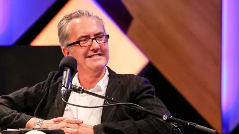 Bill Dare, wearing glasses, smiles in front of a microphone