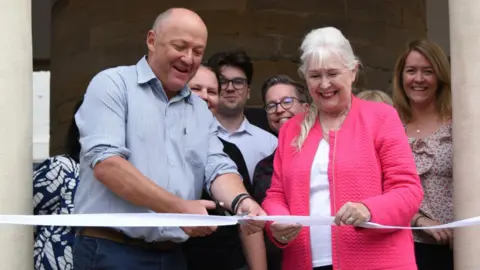 North Yorkshire Council A group of people smiling, looking at a white ribbon as a man cuts it with a pair of scissors.