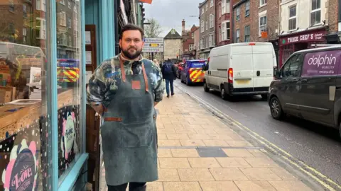 Naj Modak/BBC Jordan Thomson, owner of Love Cheese on Gillygate with queuing traffic next to him