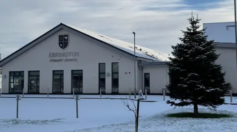 Brian Guthrie Shows a school building with the words Ebrington Primary School on it and a Christmas tree to the right with snow on the ground. 