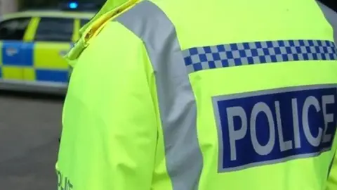 A generic photograph of a police officer wearing a high-visibility yellow jacket with the words police written in capital letters on the back. A police car is in the distance.