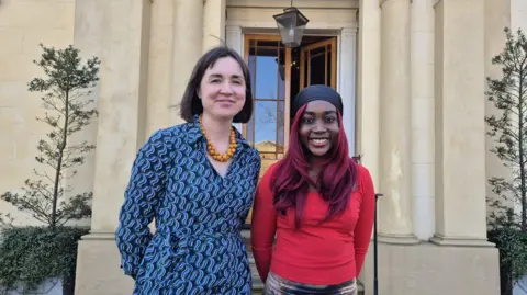 Sally and Princess stand outside the entrance to the cream-coloured stone mansion, which has columns and tall plants on either side and a blue plaque marking the author's achievements on the right