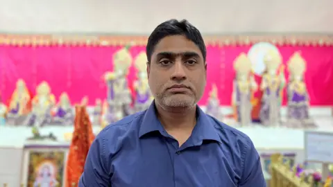 Dr Rajesh Gurrala has short black hair and a blue open-collared shirt, he is standing in front of a pink backdrop and colourful pictures and statues