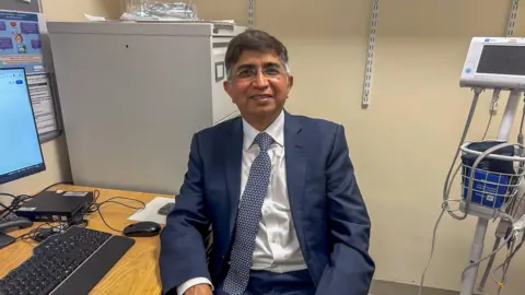 A man in a blue suit sits at a desk and smiles into the camera. 