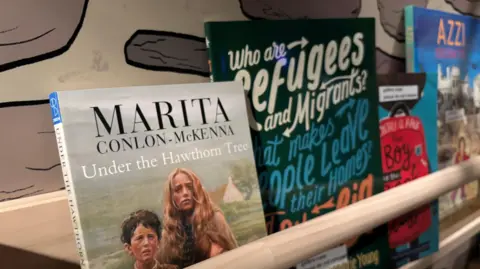 A bookshelf holds several books. The closest and biggest in frame is Marita Conlon-McKenna's Under the Hawthorn Tree. Beside it is one about Refugees and Migrants and beyond it are more books that are less visible. 