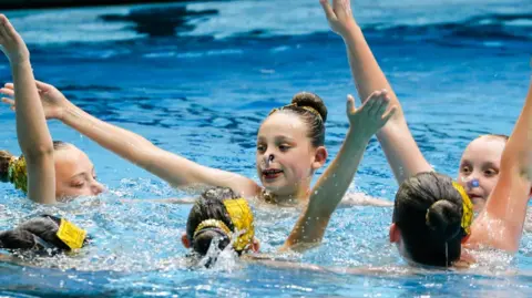 Will Johnston photography Several young swimmers hold their arms out of the water together.