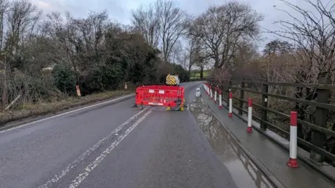 Oxfordshire County Council The A361 with a large cordoned of area in the middle. The road is above a water course, and their is a yellow and red van parked in the near distance.