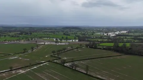 Extensive countryside is pictured from above, showing green fields and farmland bordered by hedgerows. Much of the land is covered partially by water.