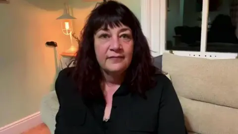 Hilary Bratton looks at the camera during a BBC interview. She has shoulder-length black hair and is wearing a black blouse.