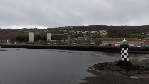 A black‑and‑white striped navigation beacon stands on the muddy shoreline of the River Clyde, with Greenock waterfront buildings and hillside housing in the background.