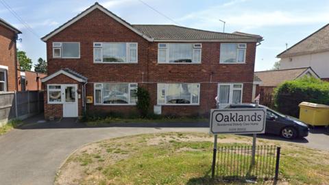 A red brick double-story building, with patch of grass in front that bears a sign reading "Oaklands"