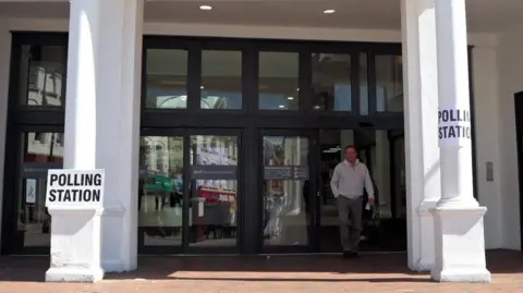 A man walking out from a large set of black doors in the front of a white building. There are two white pillars either side of the entrance with signs attached, reading 'POLLING STATION' in black lettering
