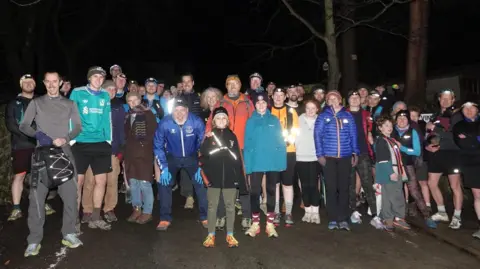 Otley Athletic Club A group of about 59 runners of all ages dressed in running gear and head torches in the dark. 