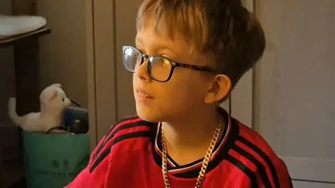 NHS Blood and Transplant A child wearing glasses sits indoors in a softly lit room, wearing a red football shirt and a gold-coloured chain necklace. Behind the child, there is a shelf with a small stuffed toy and a green container.