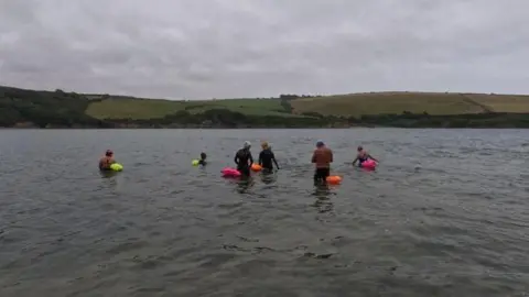 Emily Woodley Swimmers in a river with colourful floatation devices and a river bank in the distance.