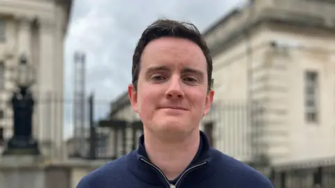 A man with dark hair and a blue zip up top is standing in front of a white stone building with black railings