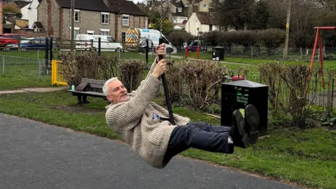 Warminster Town Council A middle-aged man with white hair and a moustache appears to enjoy riding on a zipline in a small play park. 