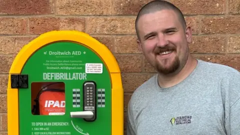 A man wearing a grey T-shirt with the words Diamond Electrical Installations written on it, is standing next to green and yellow defibrillator box that is attached to a brick wall.