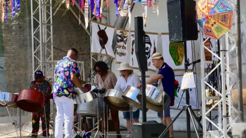 A photo of a band playing on steel drums. There are five people playing facing out towards an audience (which is not shown on the photo) and a conductor at the front. The are paintings behind them and colourful bits of fabric hanging off the front of the stage. 