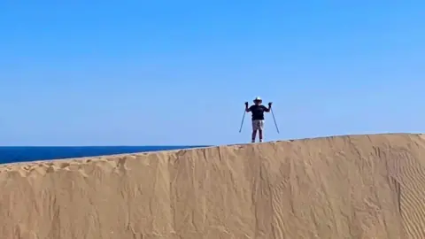 A woman in the distance trekking in sand dunes with two walking sticks wearing shorts and a hat. 