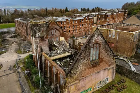 A gutted church building, built of red sandstone. There is a stone tower on the left and and an ornate Art Nouveau window. The rear wall is covered in graffiti and the roof has gone.