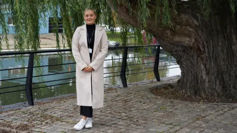 Elle Johnson is looking at the camera, wearing a long light-coloured coat, white shoes, and black top and trousers. She is smiling and standing in front of a weeping willow on a canal towpath in Ely