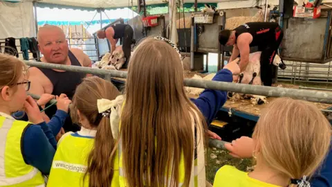 Children watch two sheep being sheared with a third shearer explaining what is happening.