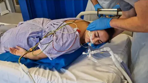 A female patient is laying on her chest, seemingly asleep, on a hospital bed. She is in a medical room with dark blue curtains separating patient beds. The patient has a medical tube attached to her nose. She is laying over a blue pillow-like device that is slowly being inflated, while a medical professional supports the patient's head.