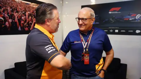 Getty Images Subramaniam Iswaran shakes hands with McLaren boss Zak Brown in front of televisions showing Formula 1