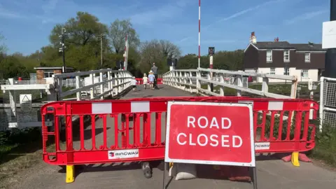 A swinge bridge over a canal that is blocked off with a sign saying 'road closed'. There are four people along the bridge, one adult man and three children. There are trees on the other side of the bridge and a brown and white building. 