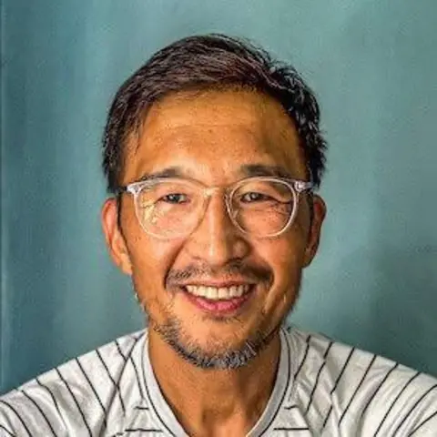 Phil Shin Uśmiechnięty mężczyzna, ubrany w T-shirt w paski i okulary. Patrzy bezpośrednio w kamerę i ma zielone tło 