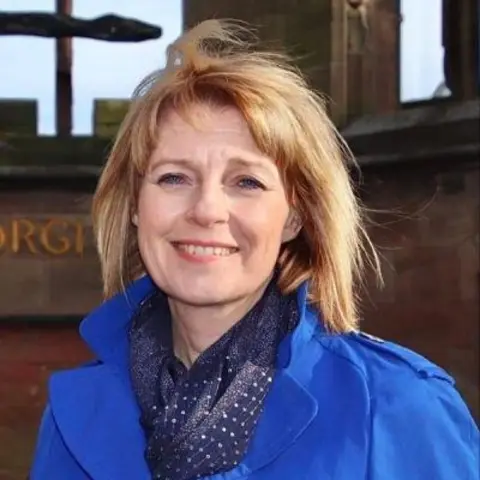 Coventry City Council A woman with reddish fair hair is wearing a blue coat and darker blue scarf. She is standing in front of the red sandstone remains of Coventry Cathedral 
